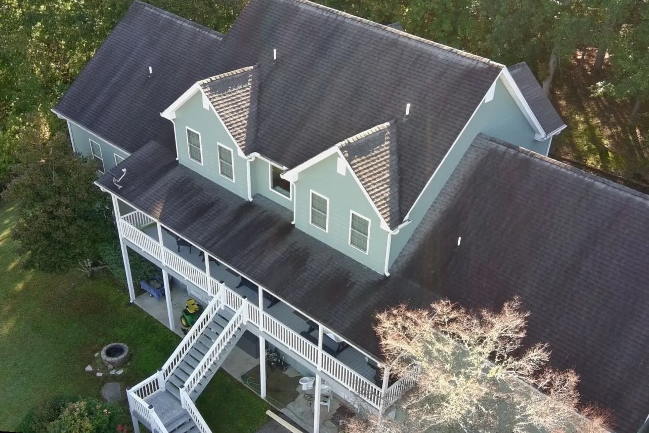 Drone view of a two-story home with dormers and a wraparound porch before replacement: a dark, stained old shingle roof