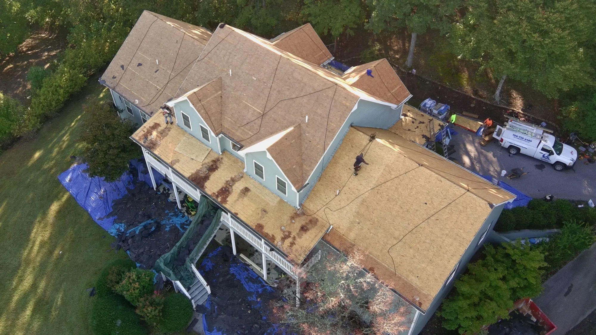 Drone view of an Iron Ridge crew tearing off an old roof down to the wood decking, with blue tarps laid over the yard and landscaping and the Iron Ridge truck in the driveway